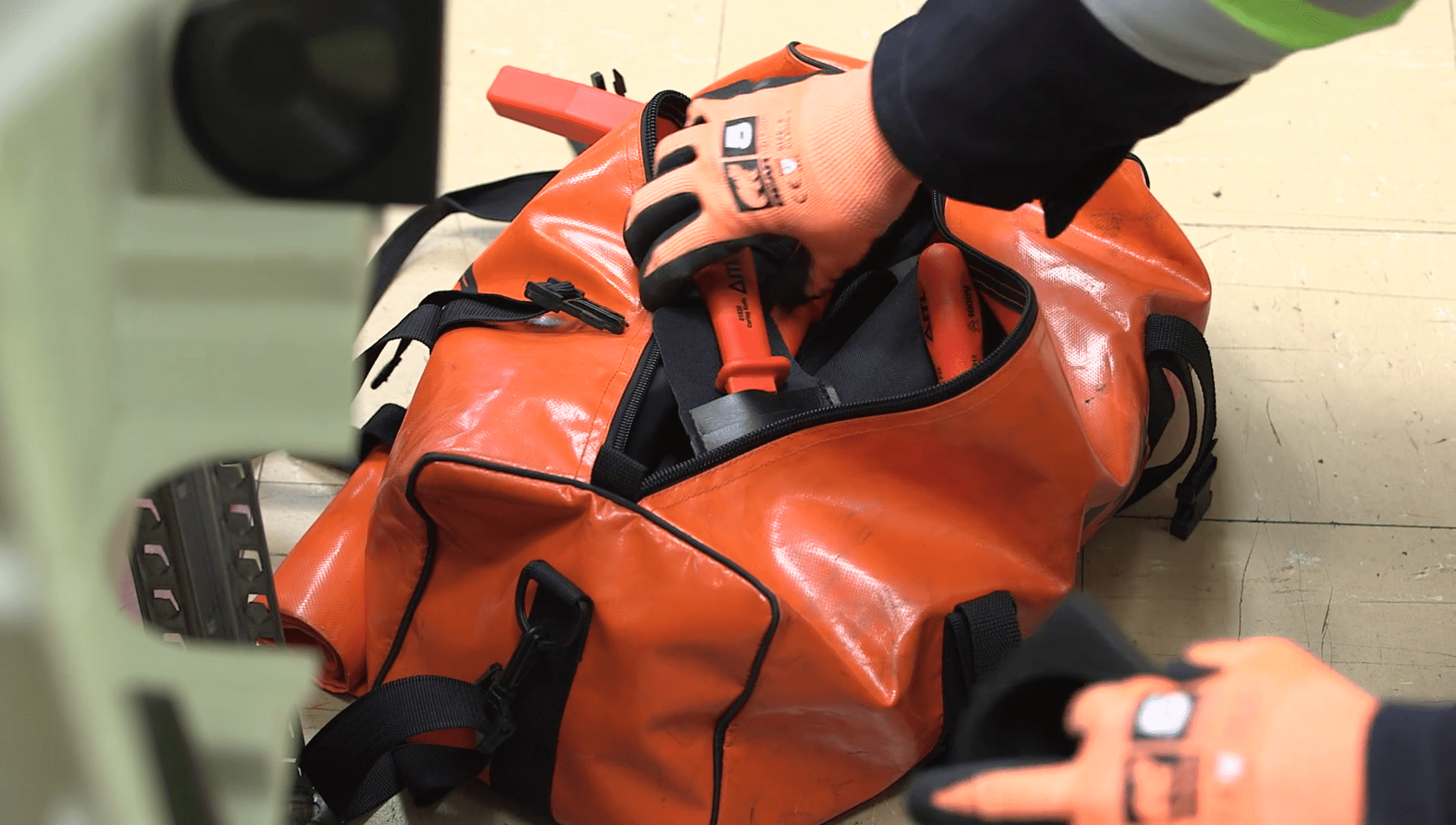 Close-up of a worker wearing an orange safety glove, placing VDE-certified insulated pliers into an open, heavy-duty orange tool storage bag.