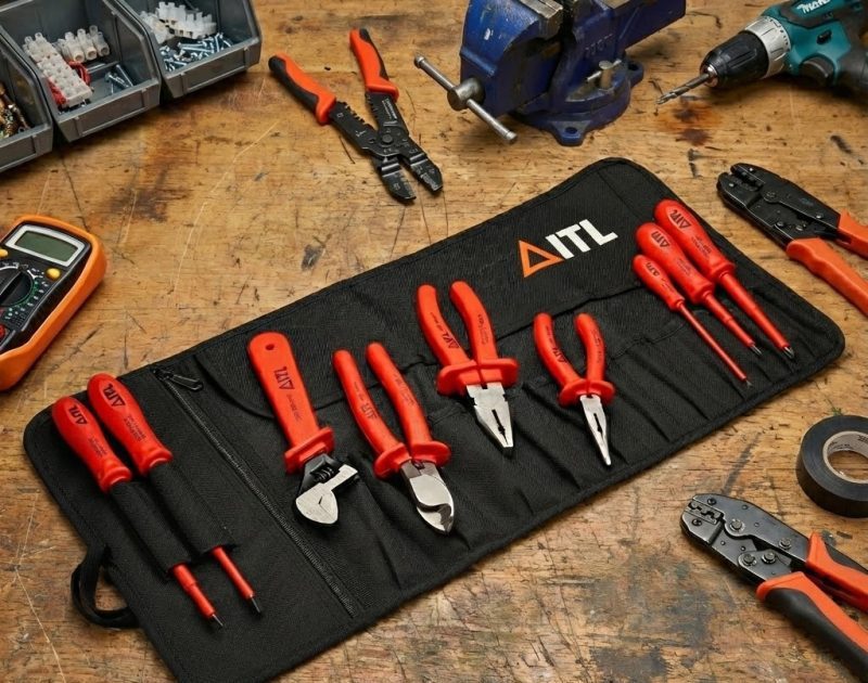 An open black canvas tool roll containing a set of orange-handled ITL insulated screwdrivers, pliers, and spanners, laid out on a wooden workbench amidst other electrical tools.
